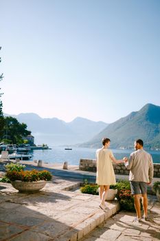 A man and a woman stand holding hands in Perast on the shore of the Bay of Kotor and look at each other