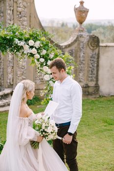 The bride reads out the wedding oath. Round wedding arch decorated with white flowers and greenery in front of an ancient Italian architecture. Wedding at an old winery villa in Tuscany, Italy.