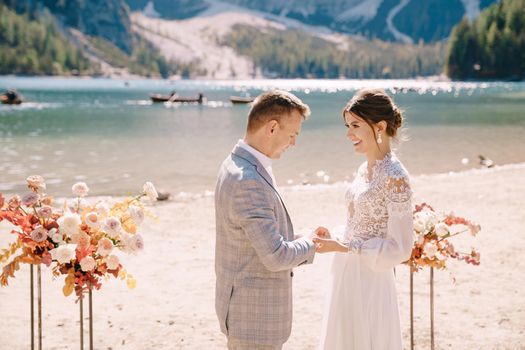 The bride puts a ring on to the groom at venue for the ceremony, with an arch of autumn flower columns, against backdrop of the Lago di Braies in Italy. Destination wedding in Europe, on Braies lake.