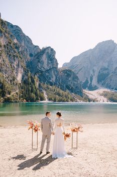 The bride and groom stand in place for the ceremony, with an arch of autumn flower columns, against the backdrop of the Lago di Braies in Italy. Destination wedding in Europe, on Braies lake.