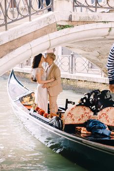 Italy wedding in Venice. A gondolier rolls a bride and groom in a classic wooden gondola along a narrow Venetian canal. Gondola floats under a stone bridge, the newlyweds kiss.