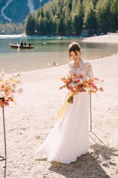 Beautiful bride in a white dress with sleeves and lace, with a yellow autumn bouquet on background of the arch for ceremony, at Lago di Braies in Italy. Destination wedding in Europe, on Braies lake.