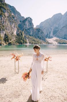 Beautiful bride in a white dress with sleeves and lace, with a yellow autumn bouquet on background of the arch for ceremony, at Lago di Braies in Italy. Destination wedding in Europe, on Braies lake.