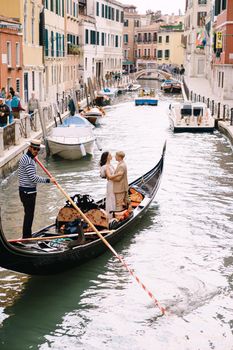 Venice, Italy - 04 october 2019: Italy wedding in Venice. A gondolier rolls a bride and groom in a classic wooden gondola along a narrow Venetian canal. Newlyweds are standing in the boat, rear view.