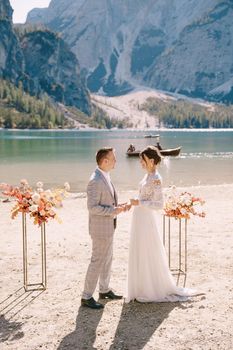 The groom puts on a ring to the bride, at the venue for ceremony, with an arch of autumn flower columns, against the backdrop of Lago di Braies in Italy. Destination wedding in Europe, on Braies lake