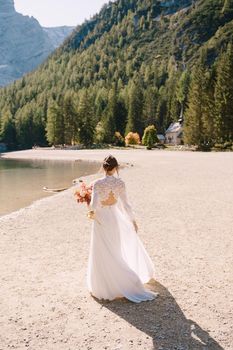 Beautiful bride in a white dress with sleeves and lace, with a yellow autumn bouquet of dried flowers and peony roses, on the Lago di Braies in Italy. Destination wedding in Europe, on Braies lake.