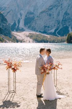 The groom puts on a ring to the bride, at the venue for ceremony, with an arch of autumn flower columns, against the backdrop of Lago di Braies in Italy. Destination wedding in Europe, on Braies lake