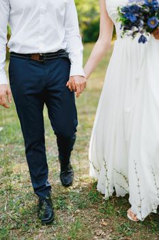 Bride with a bouquet of blue flowers and the groom walk side by side in the olive grove and hold hands. High quality photo