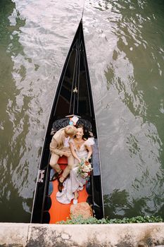 Italy wedding in Venice. A gondolier rolls a bride and groom in a classic wooden gondola along a narrow Venetian canal. Newlyweds are sitting in a boat on the background of ancient buildings.