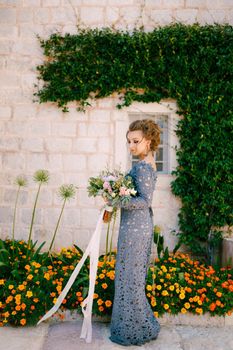 A bride in a blue dress with a bouquet stands at the wall of a house with a green liana and orange flowers, close-up