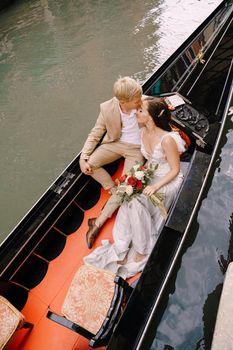 Italy wedding in Venice. A gondolier rolls a bride and groom in a classic wooden gondola along a narrow Venetian canal. Newlyweds are sitting in a boat on the background of ancient buildings.