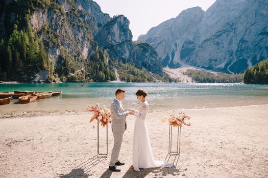 The bride puts a ring on to the groom at venue for the ceremony, with an arch of autumn flower columns, against backdrop of the Lago di Braies in Italy. Destination wedding in Europe, on Braies lake.