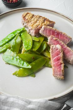 Fried tuna steak in sesame with spring onions and sugar snap peas, on plate, on white stone background