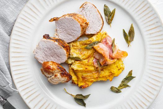 Roasted pork loin with mash potatoe gratin, sage and prosciutto, on plate dish, on white stone background, top view flat lay