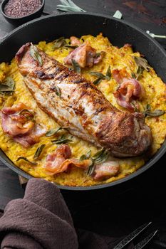 Roasted pork loin with mash potatoe gratin, sage and prosciutto, on frying cast iron pan , with barbeque knife and meat fork, on old dark wooden table background