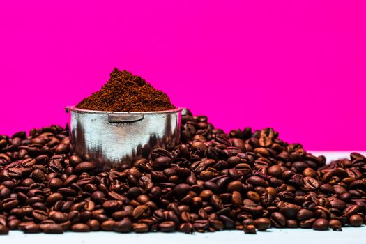 Coffee beans and portafilter with ground coffee in a composition on a pink background.