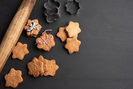 baked star shaped gingerbread cookies, wooden rolling pin and metal cutters on a black table