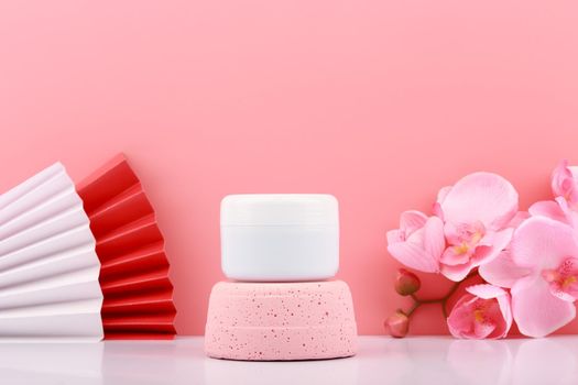 White cosmetic jar on pink pedestal against pink background with copy space decorated with wavers and flowers.