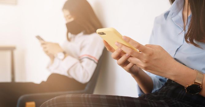 Two Asian young woman chatting in smartphone and wearing mask sitting distance protect from COVID-19 viruses for social distancing for infection risk.
