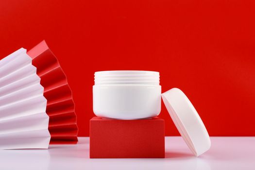 Selective focus, close up of white opened creme jar on red podium against red background decorated with wavers. 