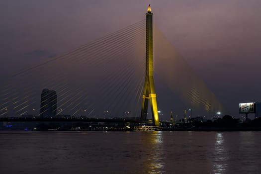 Rama VIII bridge, Rope Bridge across the Chao Phraya River, A tall V-shape pylon tower with view blue sky is background and Cityscape at night of Bangkok.