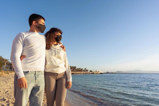 Mixed race heterosexual couple stand hugging on sea shore at beach wearing protective mask for restriction from Covid-19 pandemic looking towards the horizon at sunset. Young people trusting future