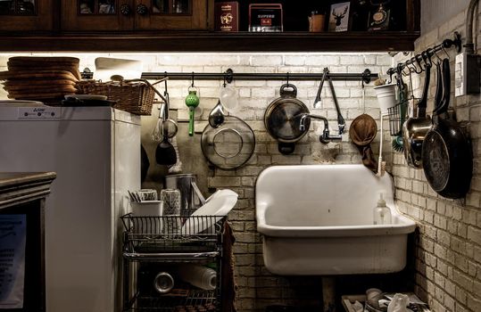 Kitchen with tools, pans, pots hanging on brick white wall and glass bottles placed on a wire rack, wooden tray placed on the freezer and ceramic tub.