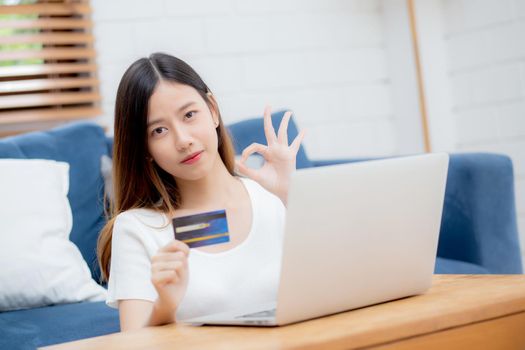 Young asian woman sit on sofa using laptop computer shopping online with credit card buying to internet, happy girl payment with e-commerce on couch, purchase and payment, business concept.