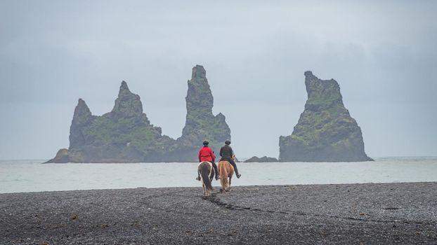 View of basalt stacks Reynisdrangar, black sand beach near Vik and two people in horseback riding, South Iceland, summer