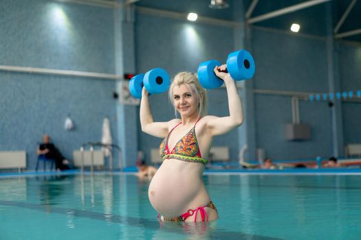 Pregnant woman standing in water with dumbbells indoors swiming pool