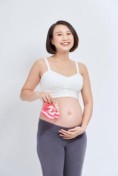 dreamy pregnant woman holding baby booties near tummy on white