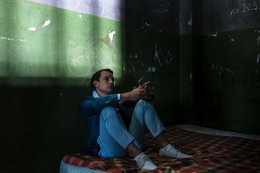 Depressed young man sitting on a mattress in a dark prison cell during custody