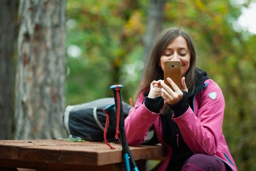 Hiker girl resting on a bench in the forest. Backpacker with pink jacket holding cell phone.