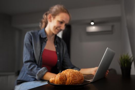 Girl working on a laptop at home. Focus on croissant.