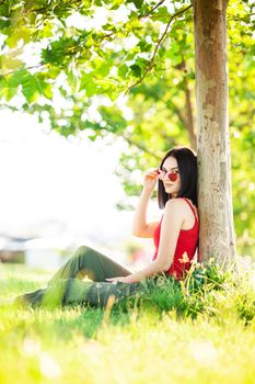 girl with dark brown hair and red sunglasses posing under a tree