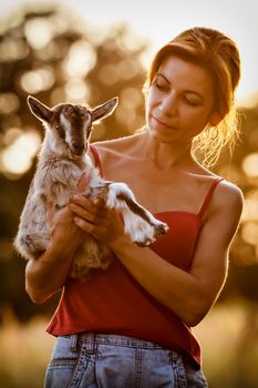 Beautiful woman with a small and nice goatling on her hands in a sunset background