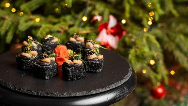 Sushi with black rice, crab meat, avocado, smoked salmon mousse, oar caviar, masago, shrimp cocktail and edible gold leaf with ginger on black table for Christmas with a Christmas tree on background.
