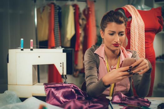 Beautiful young fashion designer sits in front of the sewing machine and using mobile phone. Vintage concept.