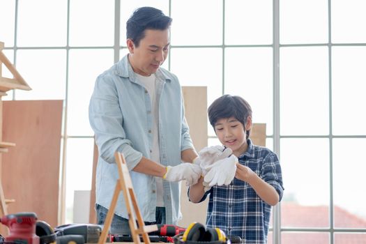 Asian father help his son to wear gloves before work with woodwork in their house. Concept of good relationship with hobby or activities in happy family.