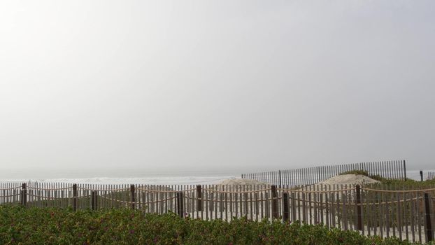 Wooden picket fence, sandy misty beach, California USA. Pacific ocean coast, fog haze on sea shore.
