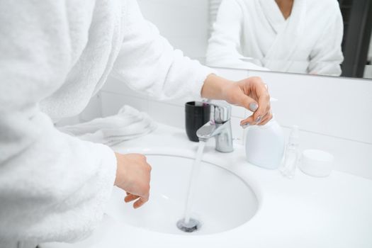  Woman in white robe standing near sink and washing hands. 