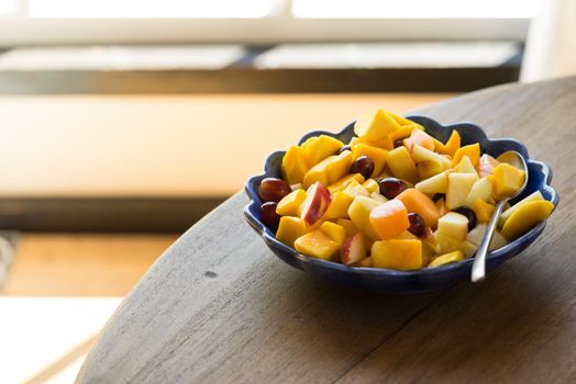 Blue bowl with fruitsalad of mango, apple, melon, grape and peach standing in the sunlight on a wooden table in front of a window in the kitchen