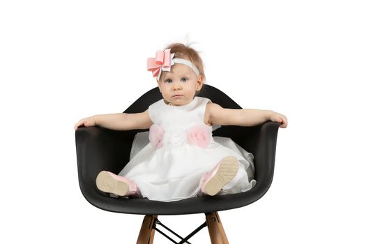 The child is one year old. Beautiful little girl sits on a chair on a white background. Funny child with a bow. Charging baby.