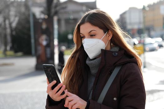 Portrait of woman wearing winter jacket with FFP2 KN95 mask using smartphone outdoors