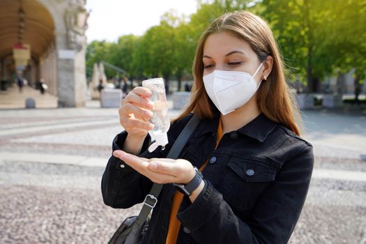 Portrait of beautiful young woman wearing KN95 FFP2 protective mask using alcohol gel sanitizing her hands in city street. Hygiene and health care concept.