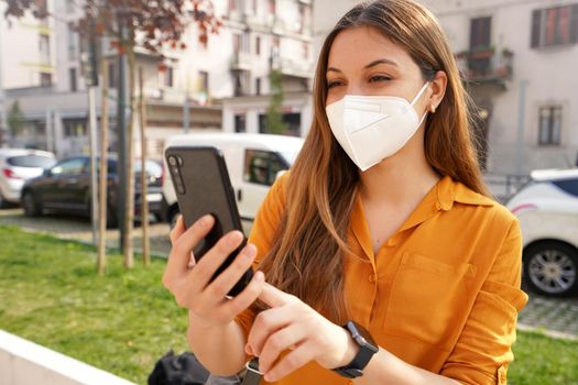 Portrait of beautiful young woman wearing KN95 FFP2 protective mask video calling with mobile phone outdoors