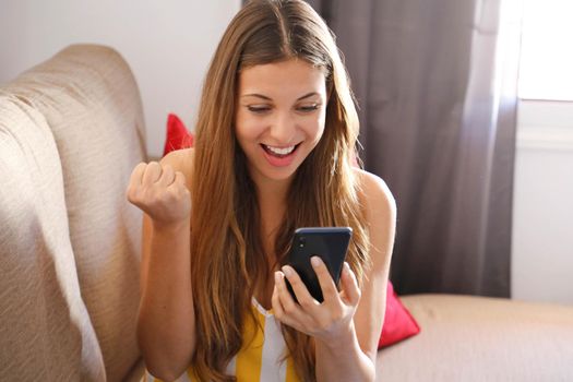 Happy overjoyed young woman holding mobile phone celebrating bid win game app victory sit on sofa at home, excited euphoric student girl winner rejoicing success with cell phone