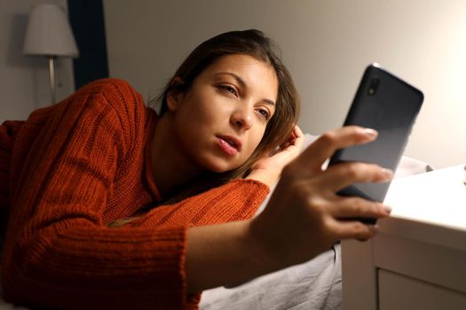 Girl suffering insomnia checks the time on the phone. Young woman awake late at night using smart phone lying in bed. Internet addiction, mobile abuse and insomnia concept.