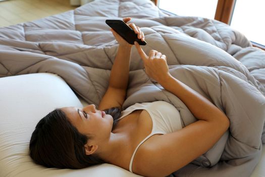 Relaxed girl using smart phone lying on the bed in the morning. Addicted to technology concept.