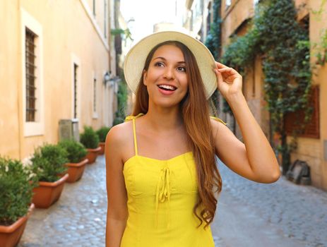 Elegant portrait of beautiful fashion woman with hat and long hair in yellow summer dress walking in Trastevere neighborhood Rome, Italy.